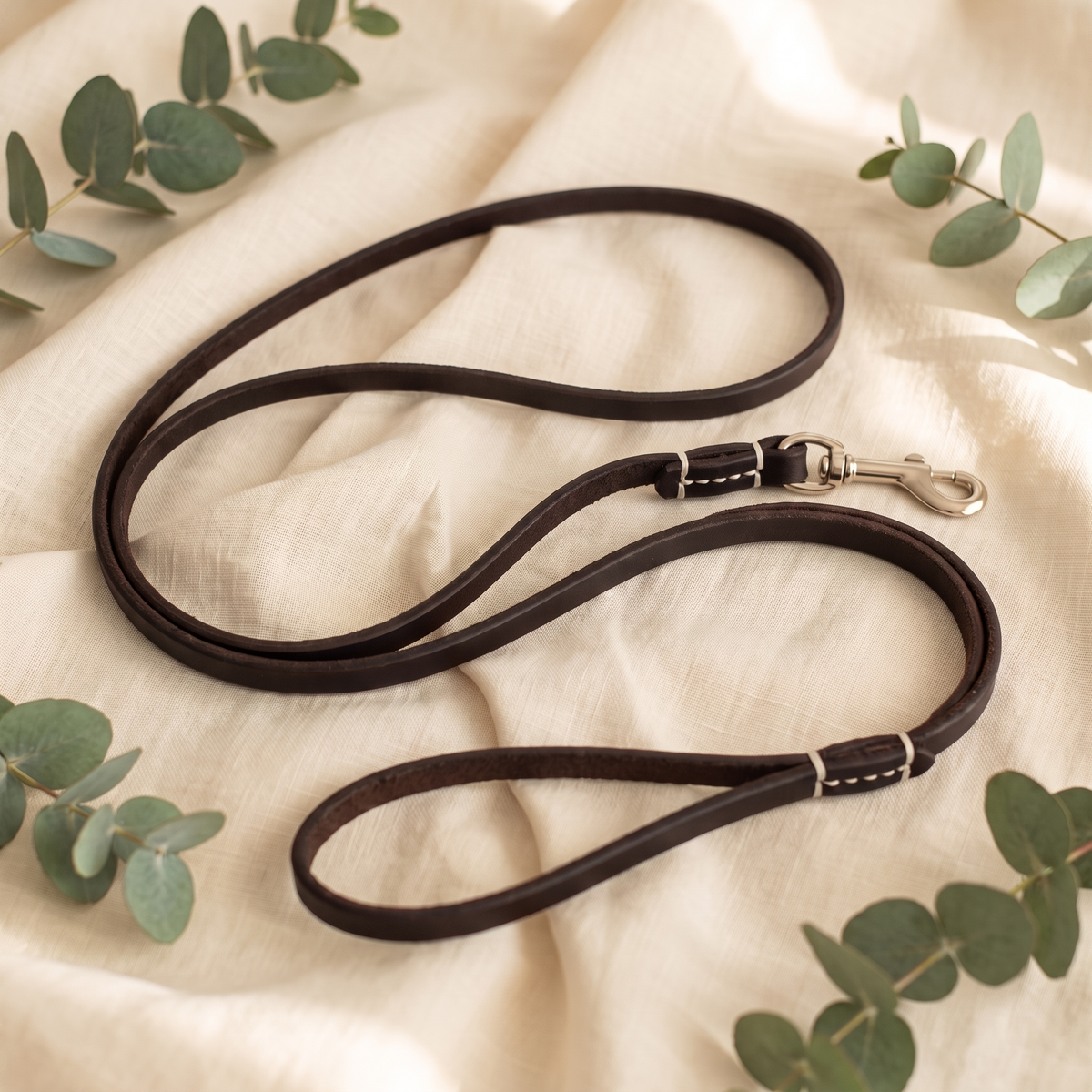 Brown Furmonogram leather dog leash, coiled on cream linen with eucalyptus