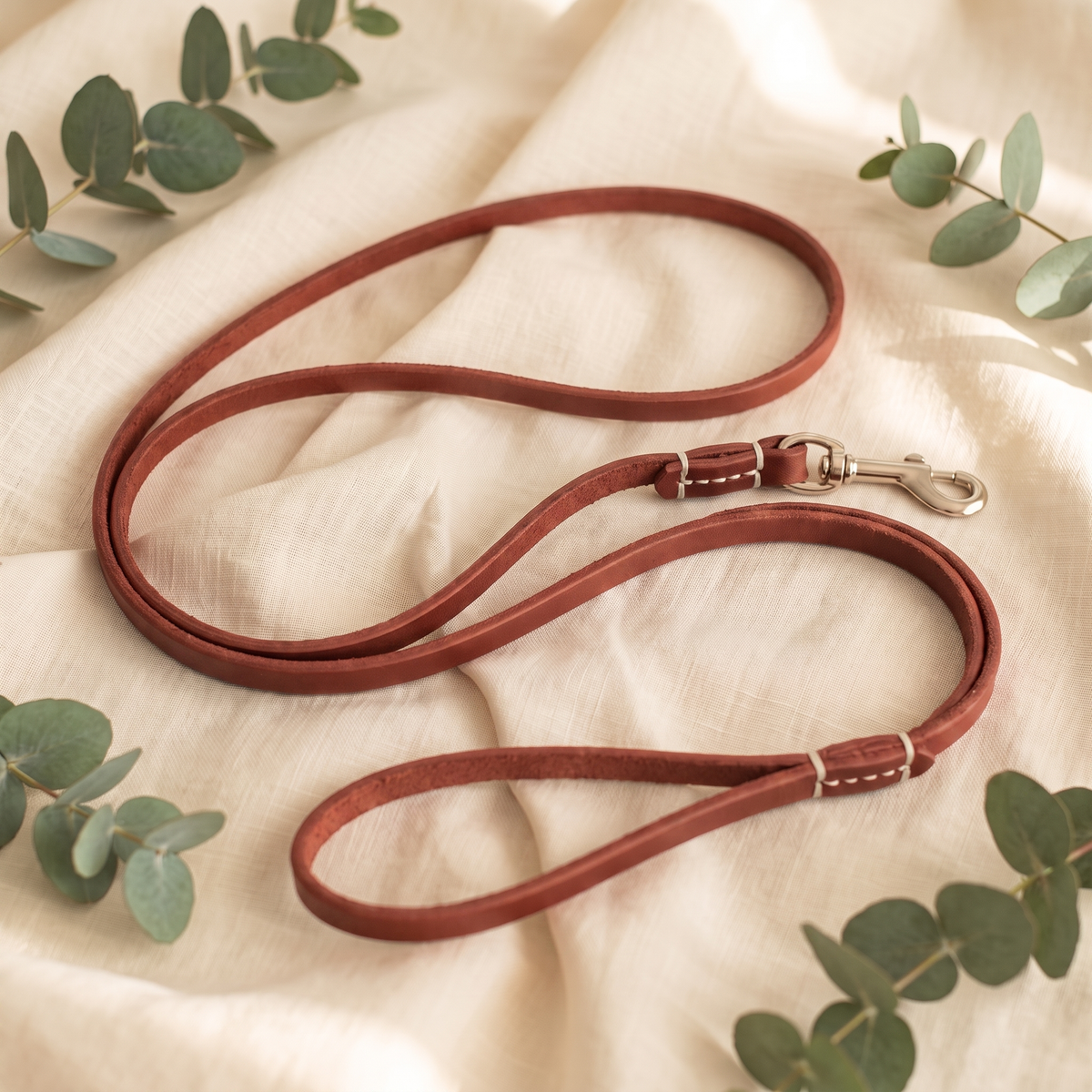 Red Furmonogram leather dog leash, coiled on cream linen with eucalyptus